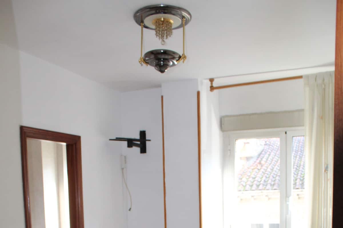 A bedroom with a bed, a wooden chair, a small wooden table, a window with curtains, and a ceiling light fixture.