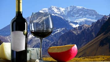 Una botella de vino, una copa de vino, un queso redondo y una manzana cortada sobre una superficie de madera con montañas al fondo.