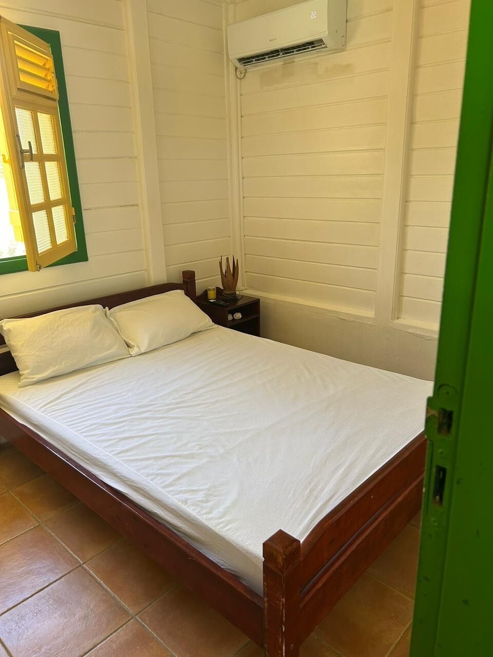 A single bed with white bedding in a room with white walls and a window with green shutters.