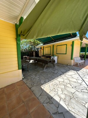 An outdoor patio with a stone floor, a wooden table, and a green awning providing shade.