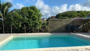En swimmingpool med en flisbelagt terrasse, omgivet af et træhegn og grøn bevoksning.