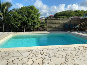 Ein Schwimmbecken mit einer terrassebelagten Steinumrandung, umgeben von einem Holzzaun und Pflanzen.