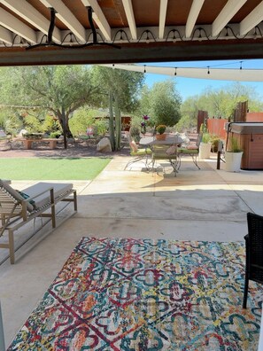 Un área de patio al aire libre con piso de concreto, un tapete colorido, una mesa metálica con sillas y una variedad de plantas y cactus.