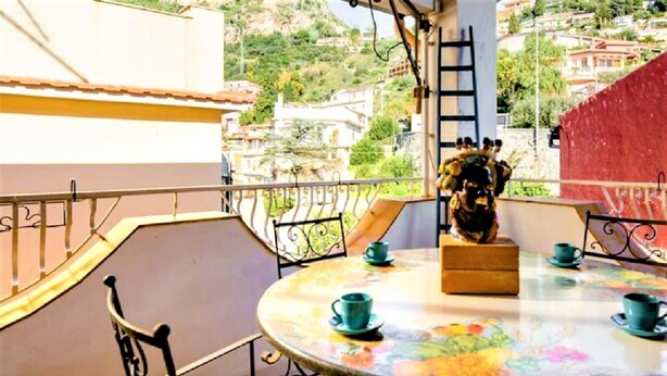 Ein runder Tisch mit einer blumigen Tischdecke, einer Statue und vier türkisfarbenen Tassen mit Untertellern auf einem Balkon mit Metallgeländer, der einen Hang mit Gebäuden überblickt.