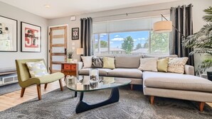 Ein modernes Wohnzimmer mit Sofa, Sessel und Couchtisch. An den Wänden hängen Bilderrahmen, ein Fenster mit Vorhängen und eine Pflanze sind ebenfalls vorhanden.