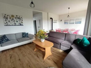 Una sala moderna con un sofá gris, una mesa de centro de madera con un jarrón de flores, y un comedor con sillas rojas y una mesa.