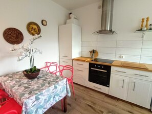 Una cocina moderna con armarios blancos, encimera de madera y horno empotrado. Hay una mesa de comedor con mantel floral y sillas rojas.