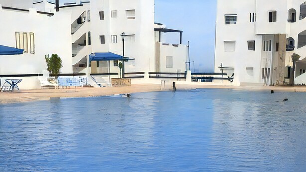 Une piscine à bordures courbes, entourée de bâtiments blancs dotés de balcons et surmontés d’auvents bleus.
