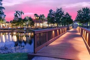 Un puente de madera con barandillas que conduce a un edificio con ventanas iluminadas, rodeado de árboles y un cuerpo de agua.