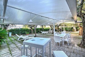 Ein Terrassenbereich im Freien mit weißen Korbmöbeln, darunter Tische und Stühle, unter einer großen Markise.