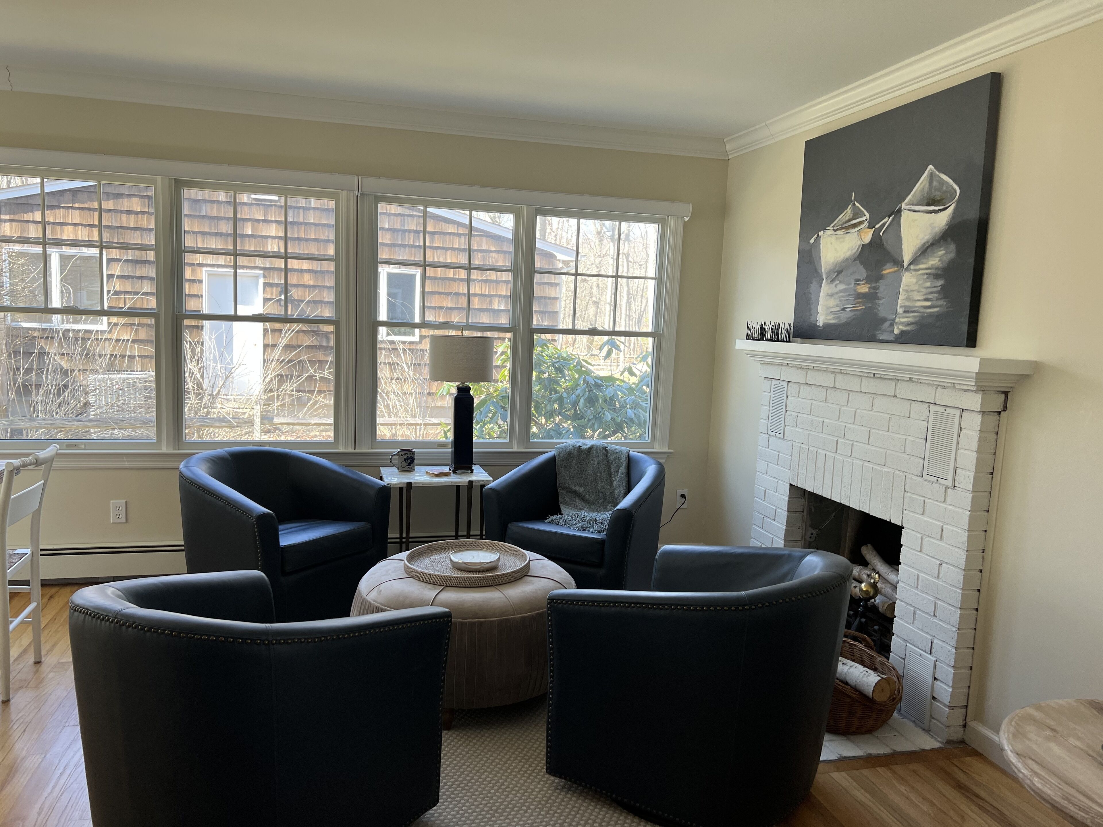 Uma sala de estar com lareira, dois poltronas, uma mesinha lateral e uma janela grande com vista para uma cerca de madeira e árvores.