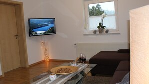 A living room with a TV, a window with a plant, a sofa, and a coffee table with snacks and a wine glass. - Small vacation apartment (65m²) with "beautiful view" Garden with playground (Nieder-Werbe)