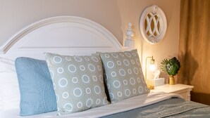 Una cama bien hecha con almohadones estampados, un cabecero blanco y una mesita de noche con una lámpara y una pequeña planta.