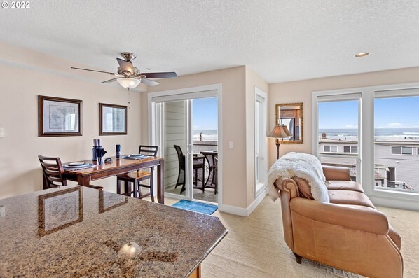 A spacious living room with a large window offering a view of the beach, a dining area with a table and chairs, and a cozy seating area with a fur throw.