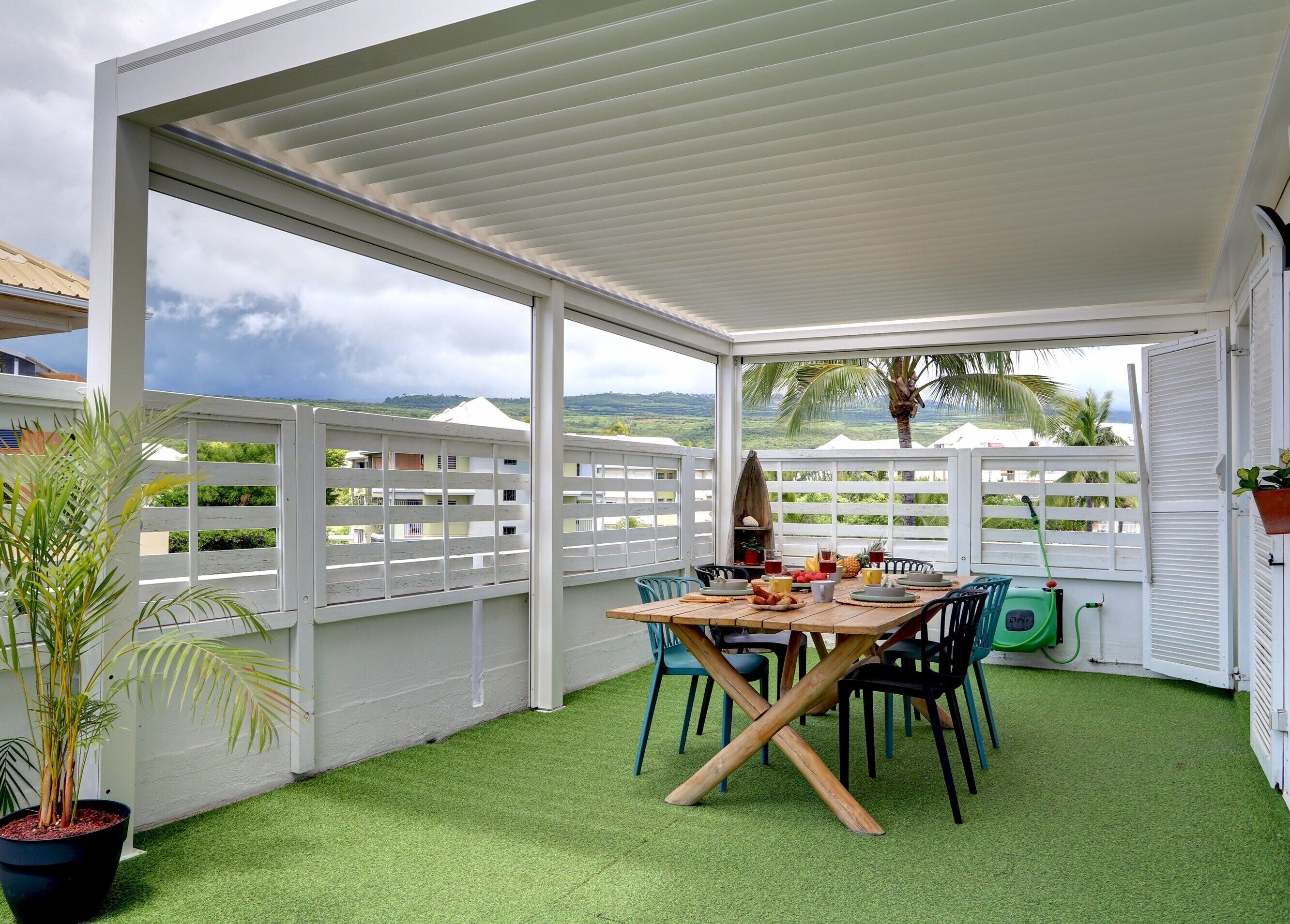 A covered patio with a table set for dining, surrounded by chairs, and a potted plant.
