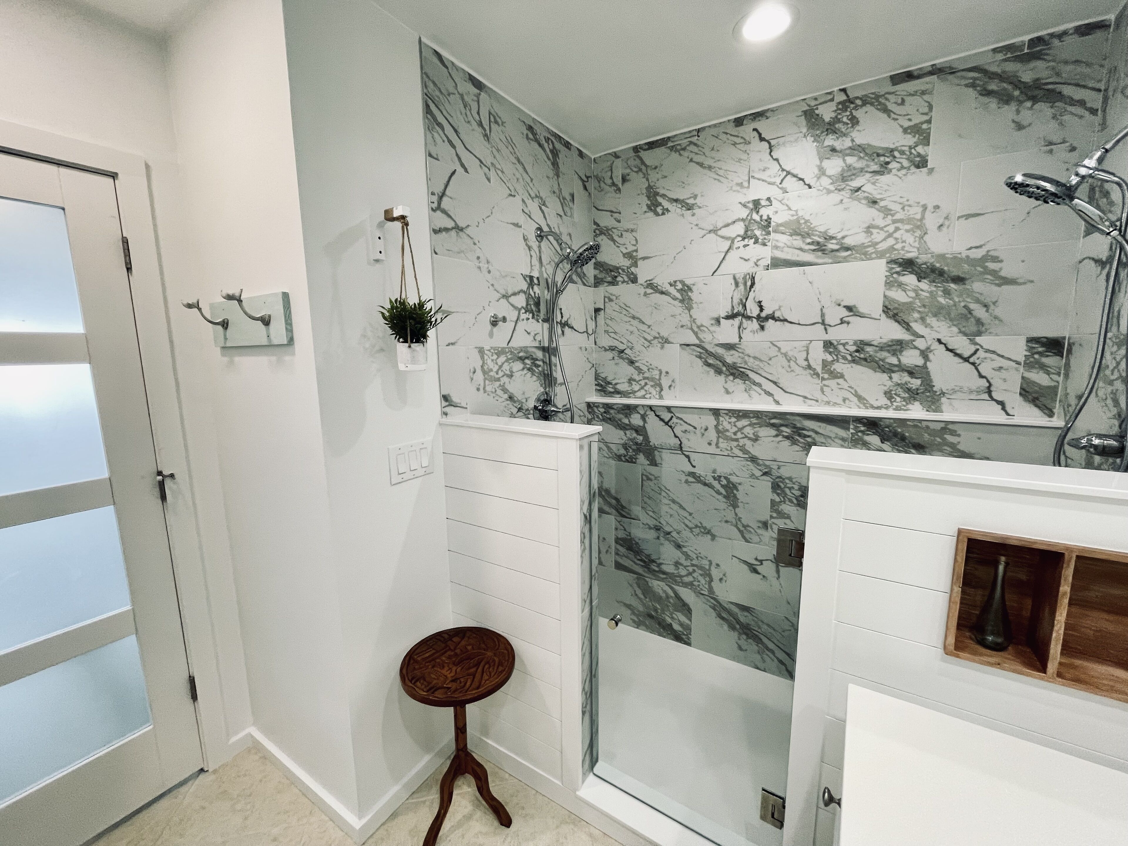 Un baño con cabina de ducha de vidrio, azulejos de mármol en la pared, un taburete de madera y un estante de madera con un jarrón.