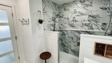Un baño con cabina de ducha de vidrio, azulejos de mármol en la pared, un taburete de madera y un estante de madera con un jarrón.