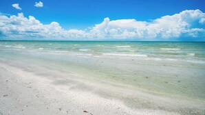 En strand med klart vand, hvidt sand og en blå himmel med skyer.