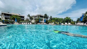 En klar swimmingpool med liggestole og parasoller, omgivet af bygninger og palmer.
