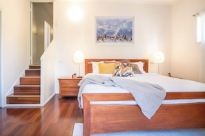 A bedroom with a wooden bed, a nightstand, a bedside table, a painting of a snowy mountain, and a staircase leading to another room.