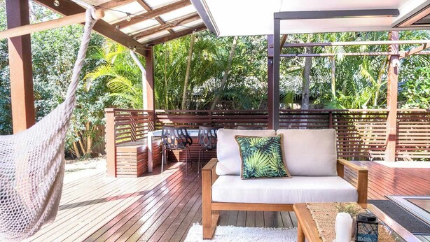A wooden deck with a hammock, a sofa with a tropical pillow, and a table with a candle and lantern. - Mid-Century Modern Retro Beach House (Caloundra Region)