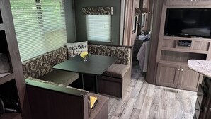 Interior compacto de una caravana con comedor, un banco de asientos, una pequeña mesa con una flor y un televisor integrado.