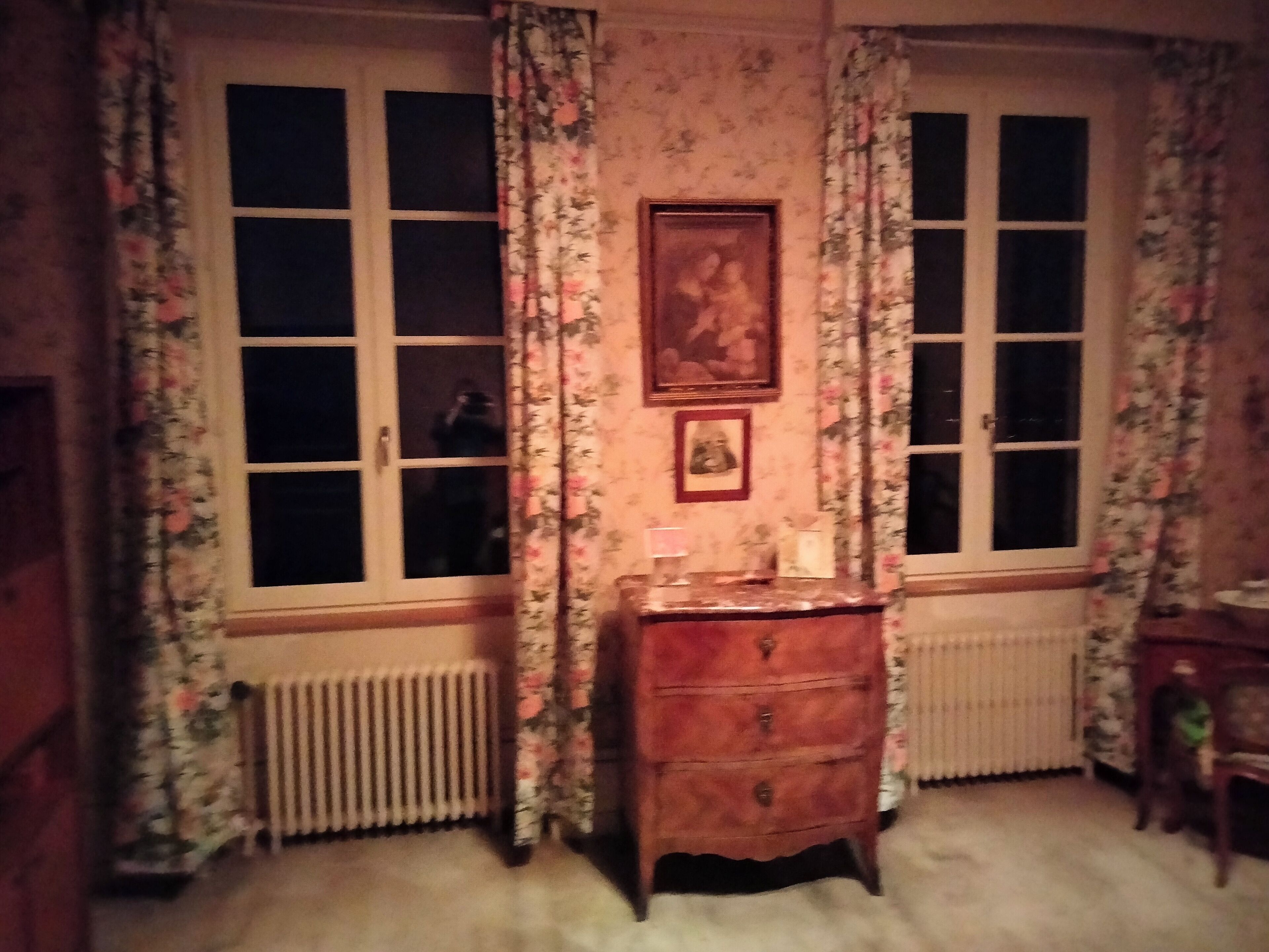 Une pièce avec deux fenêtres, des rideaux à motifs floraux, un radiateur, une commode en bois et un tableau encadré accroché au mur.