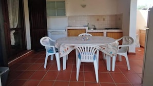 A dining area with a round table and six white chairs, a kitchen with white cabinets, and a tiled floor. - Sardinia Villasimius coastline, ins (Quartu Sant'Elena)