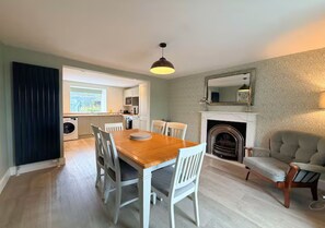 A dining area with a wooden table and chairs, a kitchen in the background, and a fireplace with a mirror above it. - Spacious 3BR Cottage in Beautiful Rural Setting - Pass the Keys (Castle Douglas)