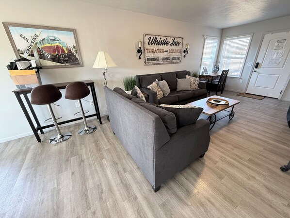 A living room with a gray sofa, two bar stools, a coffee table, a painting of a train, and a sign that reads 'Whistle Inn Theater and Lounge.'