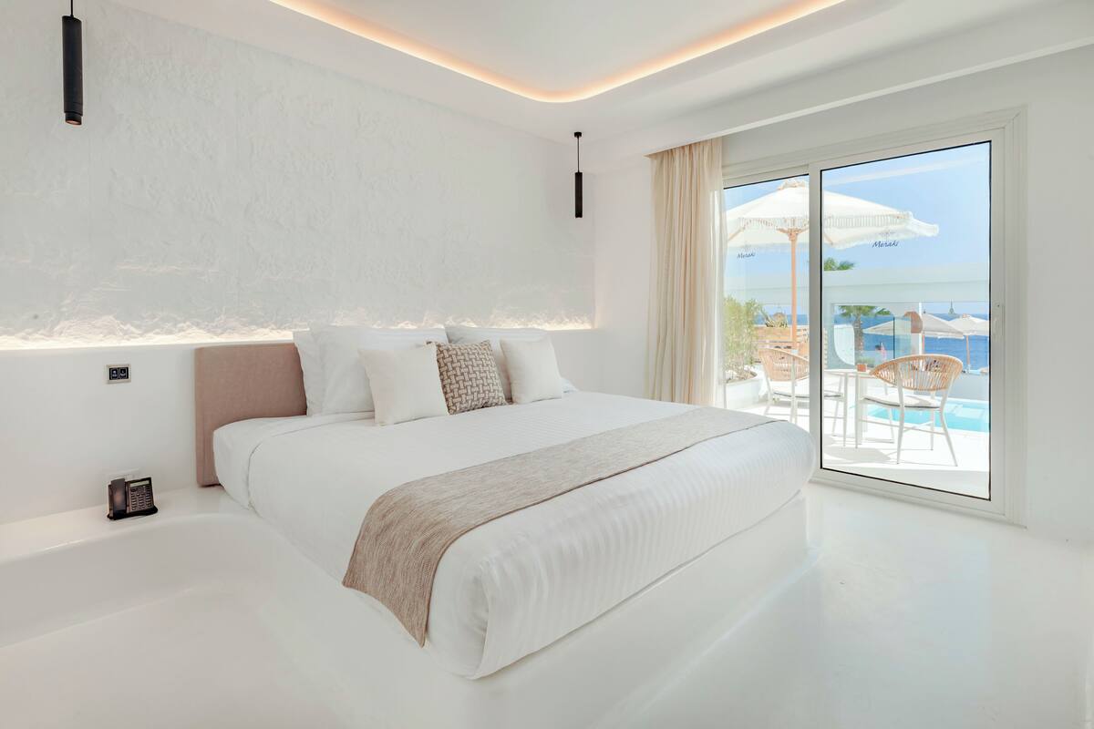 A modern hotel room with a large bed, a bedside table, and a view of a pool and outdoor seating area.