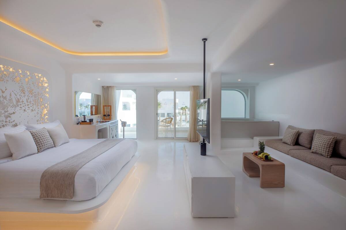A modern hotel room with a large bed, a sofa, a coffee table, and a view of the outdoors.