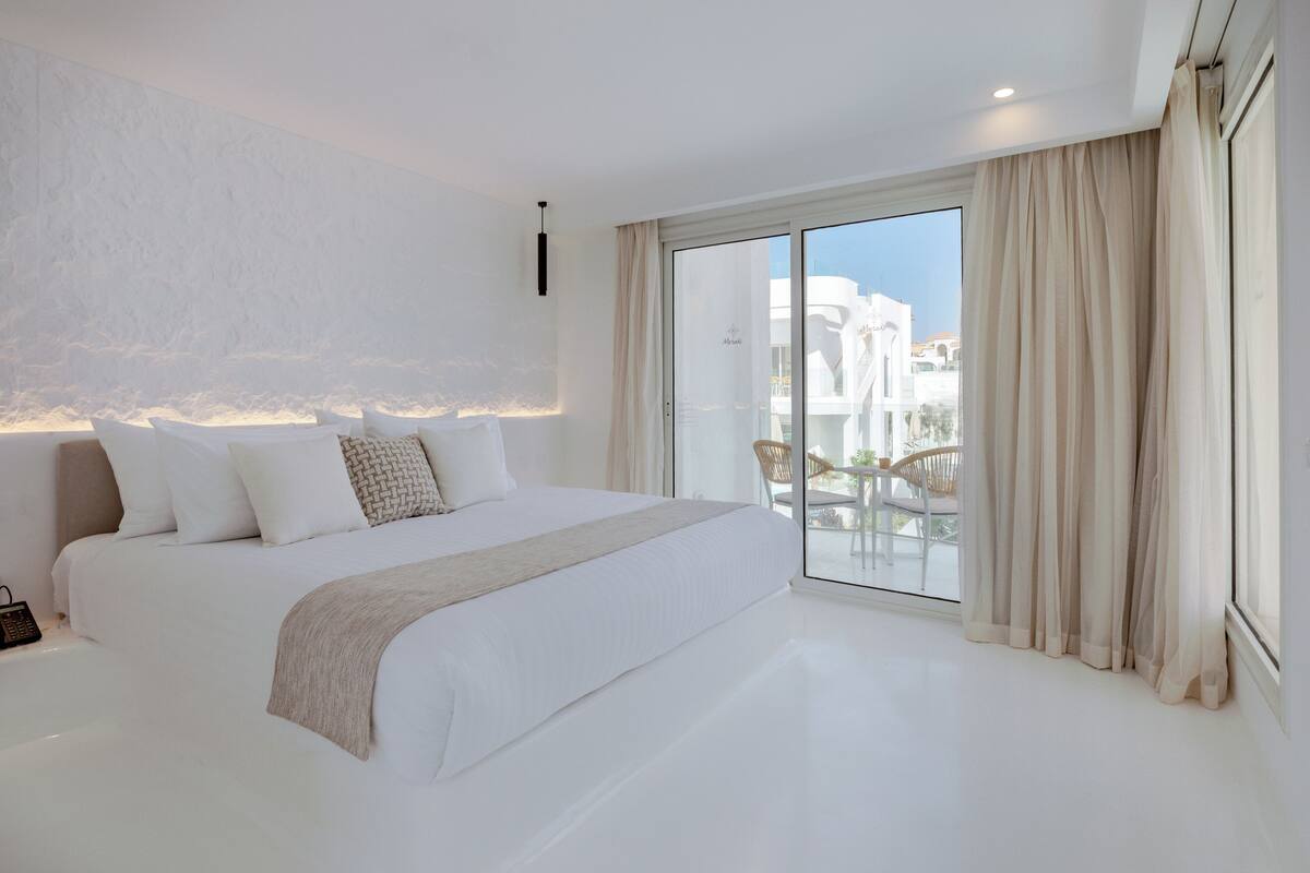 A modern bedroom with a large bed, a bedside phone, and a view of the outdoors through a sliding glass door.