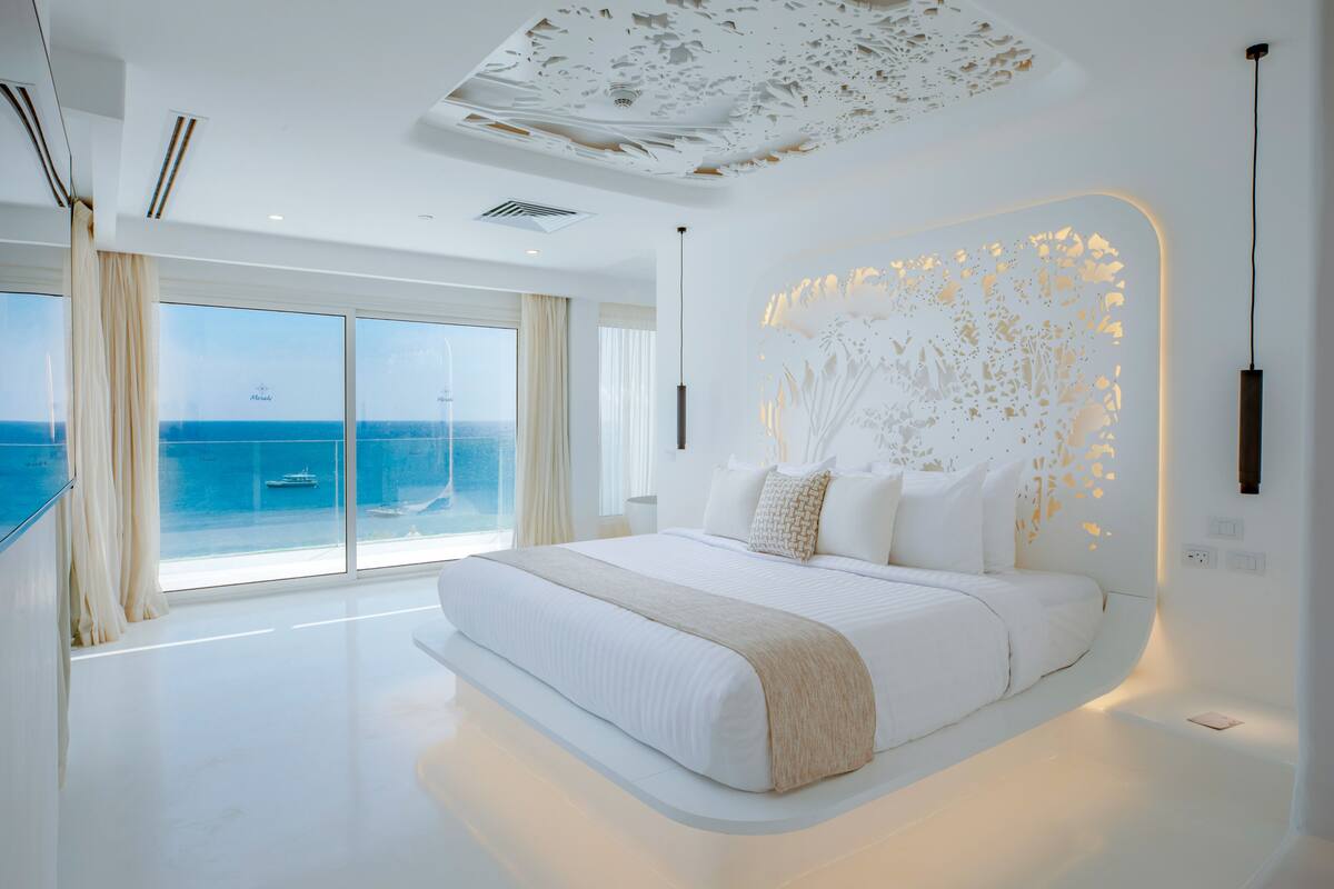 A modern bedroom with a large bed, a view of the sea, and a decorative ceiling.