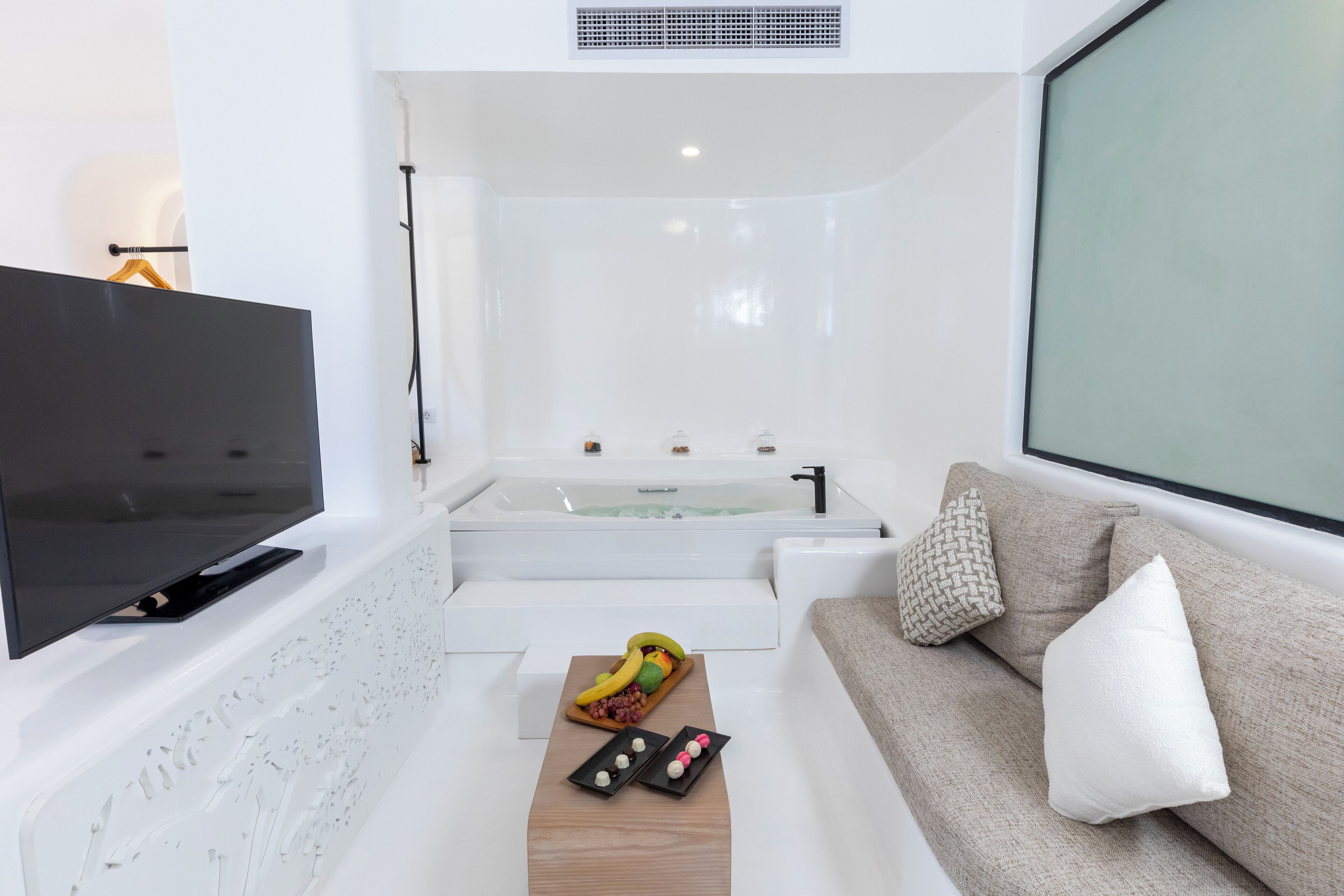 A modern living room with a flat-screen TV, a sofa with cushions, a wooden coffee table with decorative items, and a large window.