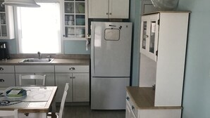 Eine kleine Küche mit Spüle, Kühlschrank und Schränken. Ein Esstisch mit Stühlen und eine Couch mit gemustertem Kissen sind ebenfalls vorhanden.