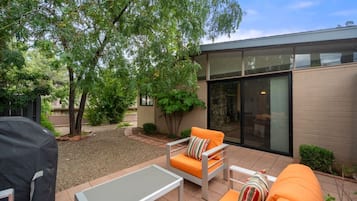An outdoor patio with a modern design, featuring a glass table, two orange sofas, and a barbecue grill.