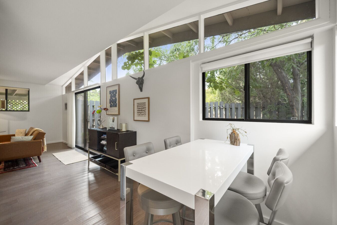 Uma sala de estar moderna com uma mesa de jantar branca, cadeiras cinzentas, um sofá marrom e um armário de madeira escura.