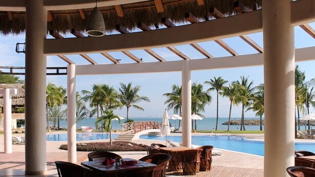 A resort pool area with lounge chairs and tables, a thatched roof, and a view of the ocean.