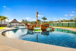 A swimming pool with two wooden ship-shaped structures, a thatched-roof building, and palm trees.