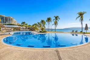 Una piscina amplia y transparente con sillas de descanso y palmeras, rodeada por un edificio de resort.