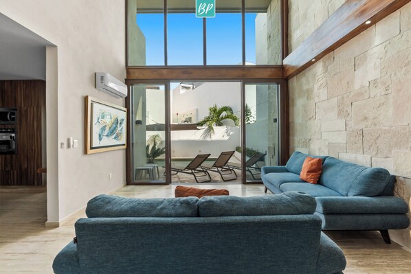 A modern living room with a blue sofa, a coffee table, and a large sliding glass door leading to a balcony with lounge chairs.