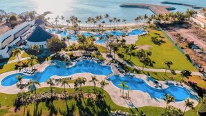 An aerial view of a resort with multiple swimming pools, palm trees, and a beach.