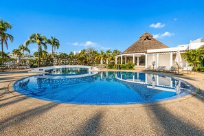 A resort pool with a circular design, palm trees, and a thatched-roof building.