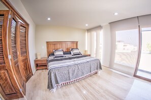 Un dormitorio con cabecera de madera, cama con manta a rayas, una mesita de noche y un ventanal con vistas a edificios.