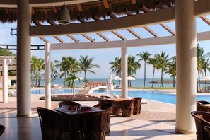 A resort pool area with lounge chairs and tables, a thatched roof, and a view of the ocean.