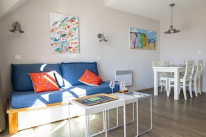 Uma sala de estar moderna com sofá, mesa de jantar e cadeiras. Há duas pinturas nas paredes e uma luminária no teto.