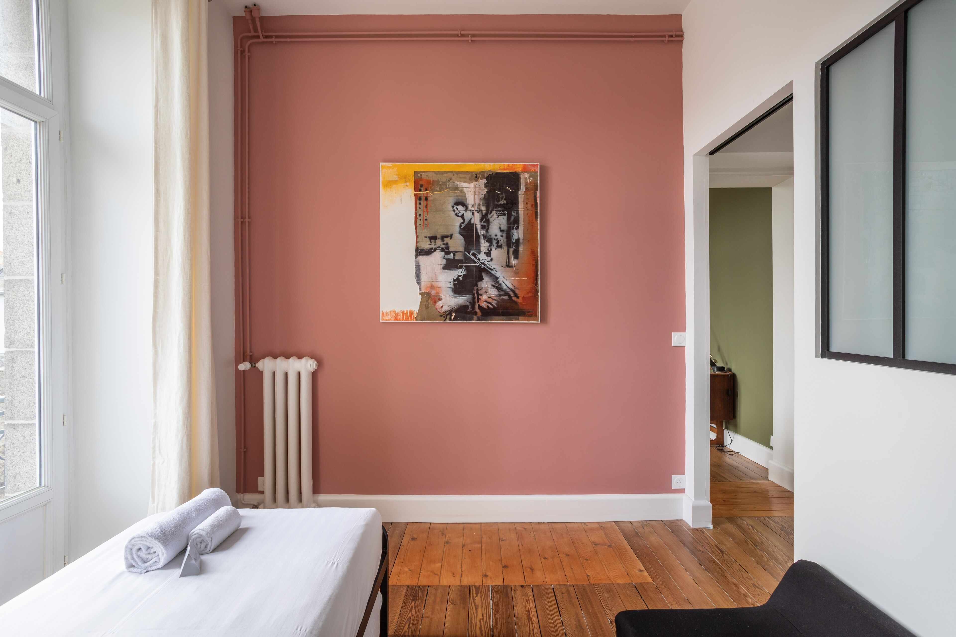 A hotel room with a bed, a radiator, a painting on the wall, and a view of the hallway.