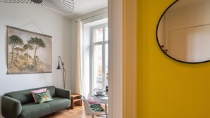 Une pièce avec un canapé vert, une petite table et une chaise avec un coussin vert. Il y a un tableau au mur et une fenêtre avec des rideaux.