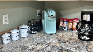 Une cuisine avec des meubles de couleur bois clair, un plan de travail en granit, une machine à café et des récipients étiquetés pour le sucre, le thé et le café.